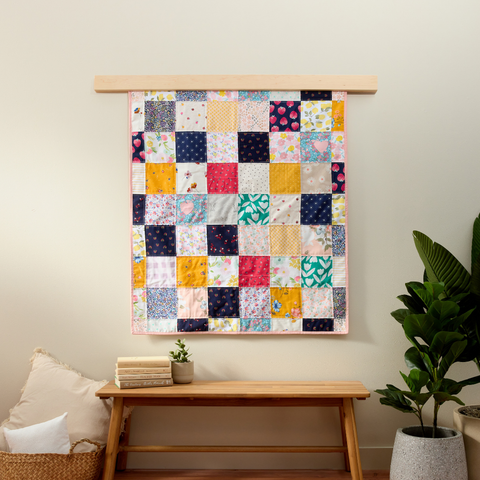 Colorful patchwork baby clothes memory quilt displayed on a wooden wall hanger above a bench, surrounded by neutral décor and green plants.