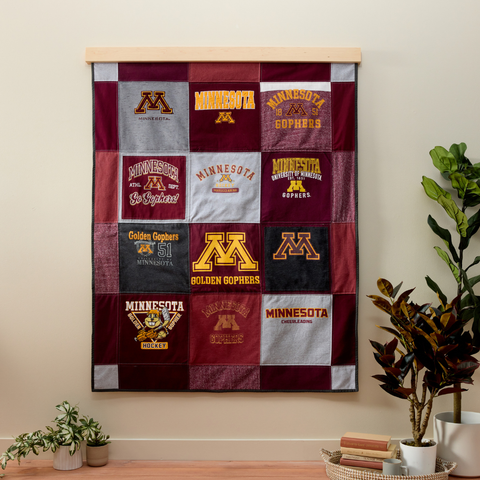 Collegiate-themed memory quilt made from maroon, gold, gray, and black shirts, displayed on a wooden wall hanger beside green plants.