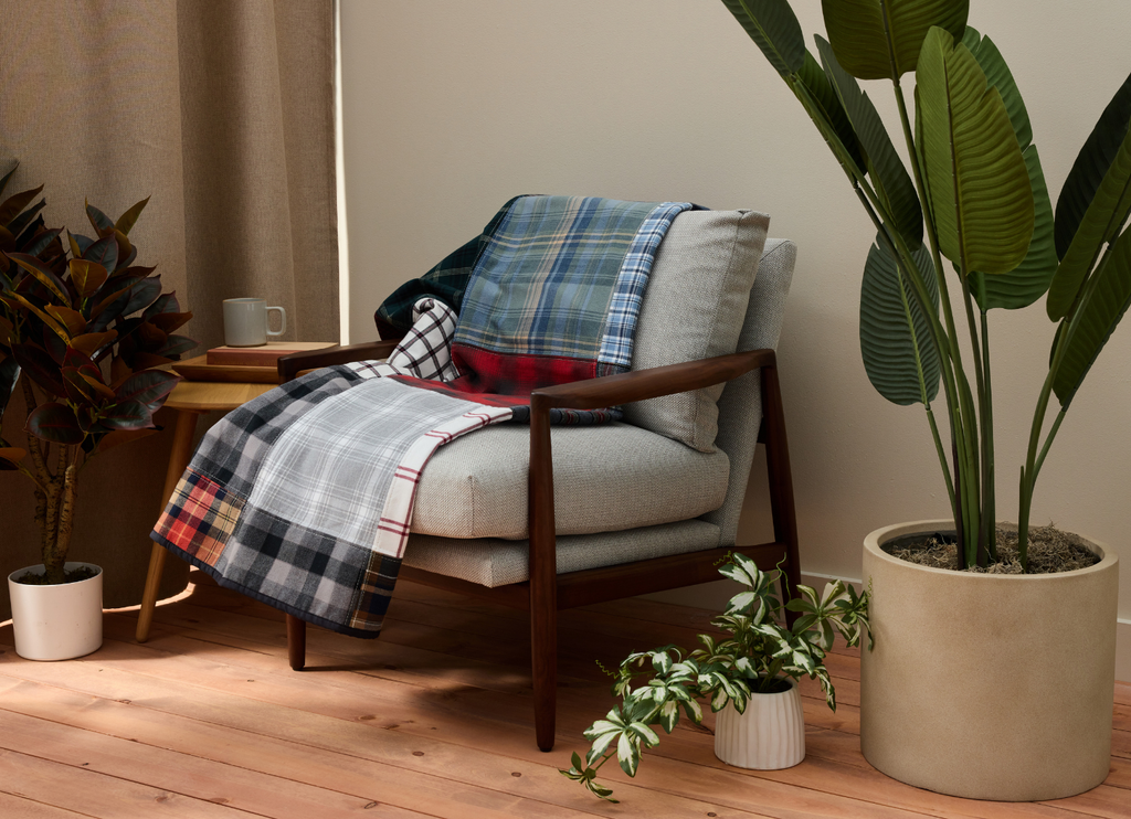 Nice Blanket Signature Memory Quilt made from plaid shirts in blue, red, gray, black, and white, draped over a cushioned wooden chair in a warm, plant-filled room.