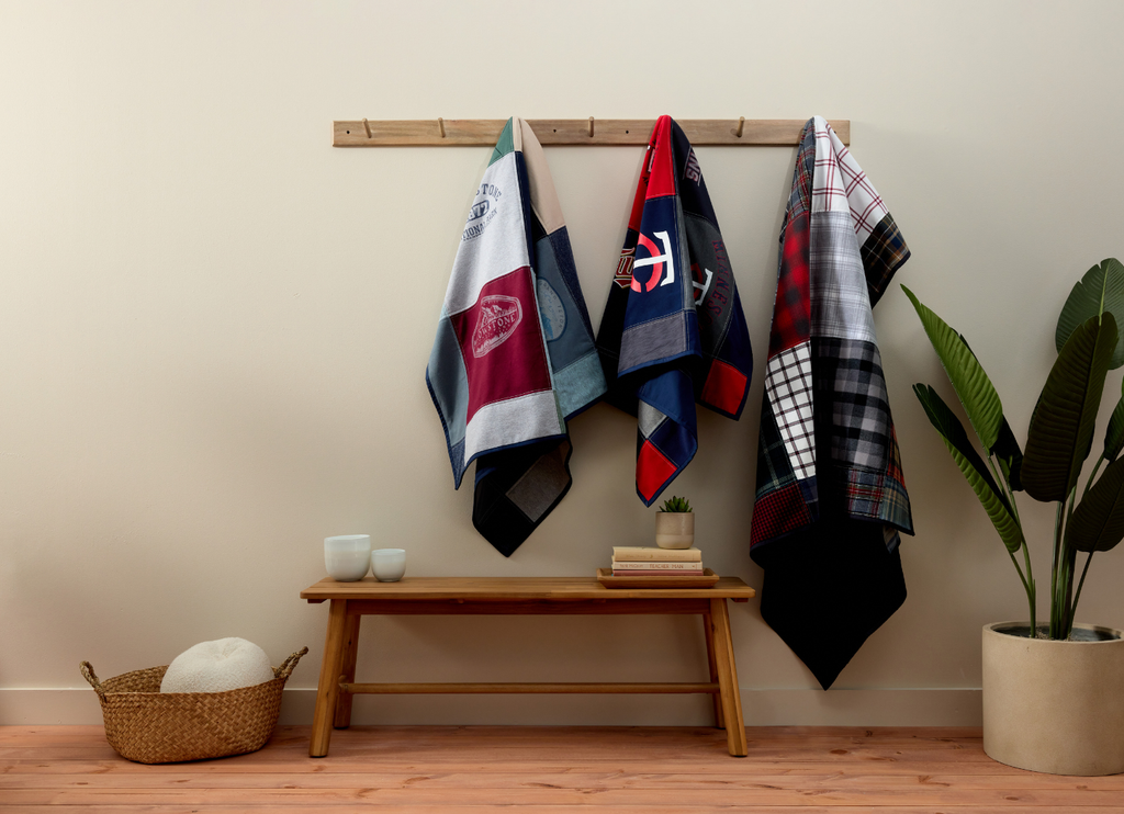 Three Nice Blanket Signature Memory Quilts made from shirts, displayed on wooden wall hooks above a bench: a travel-themed quilt, a sports-themed quilt, and a plaid shirt quilt.