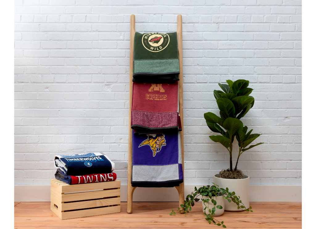 Nice Blanket Signature Memory Quilts made from Minnesota sports shirts, featuring the Wild, Gophers, Vikings, Timberwolves, and Twins, folded on a wooden ladder and stacked on a crate.