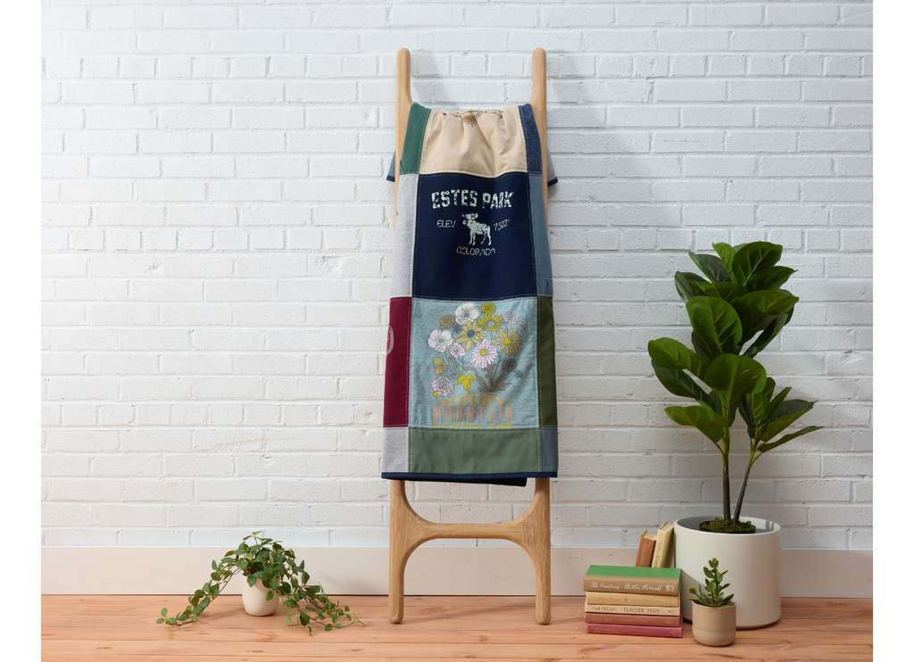 Colorful memory quilt on a wooden blanket ladder against a white brick wall with plants and books on a wooden floor.