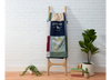 Colorful memory quilt on a wooden blanket ladder against a white brick wall with plants and books on a wooden floor.