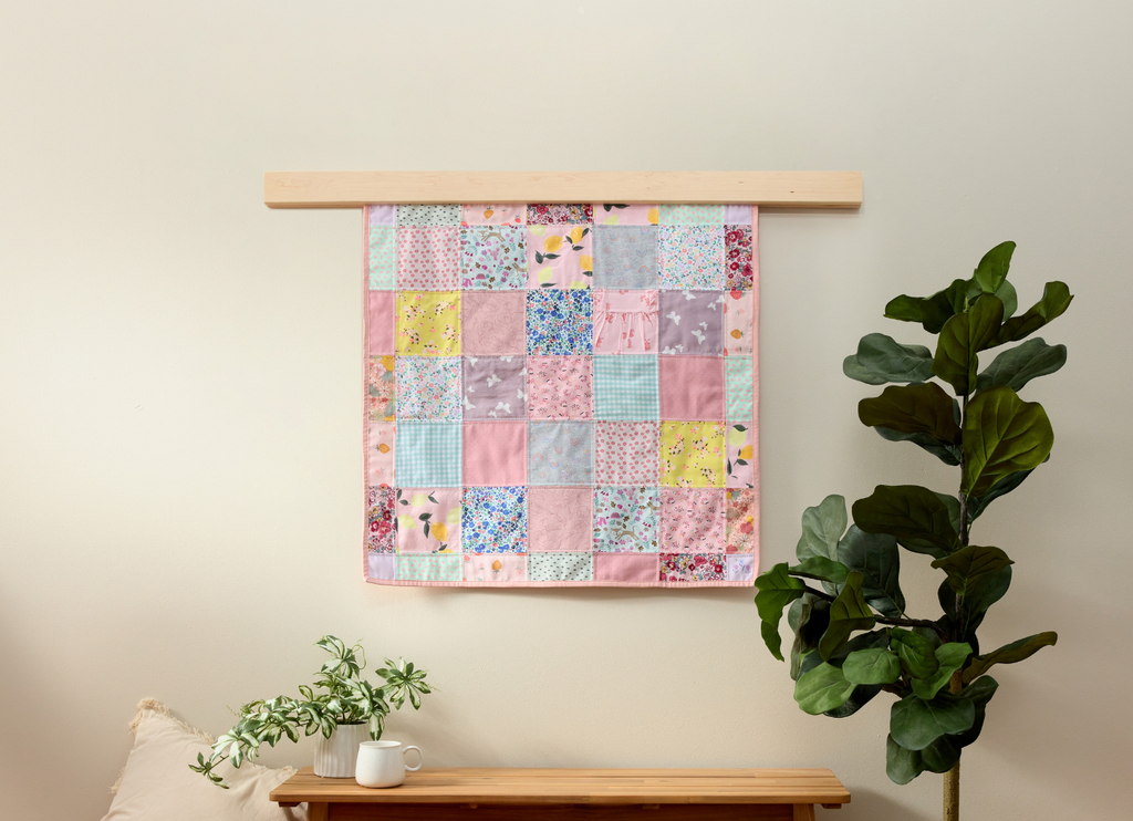 Patchwork memory quilt hanging on a wall with a plant and bench below.
