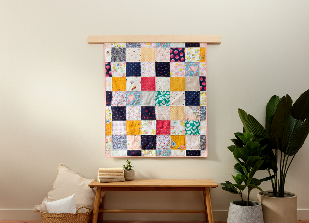 Nice Blanket Little Memories Quilt made from colorful infant and toddler clothes, displayed as wall art above a wooden bench in a warm, neutral room.