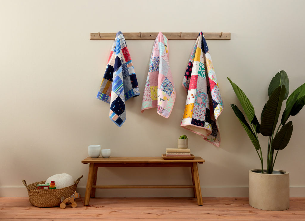 Colorful Little Memories memory quilts hanging on a rack against a plain wall with a wooden bench and plant in the foreground.