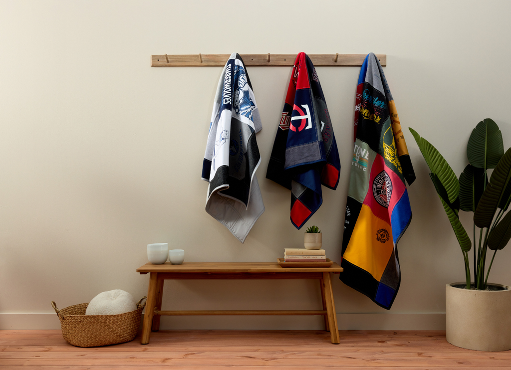 Colorful memory quilts hanging on a rack against a plain wall with a wooden bench and decor items.