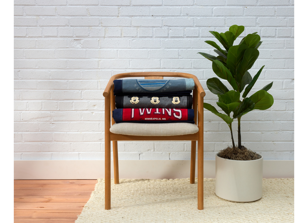 Wooden chair with folded memory quilts on it, next to a potted plant against a white brick wall.